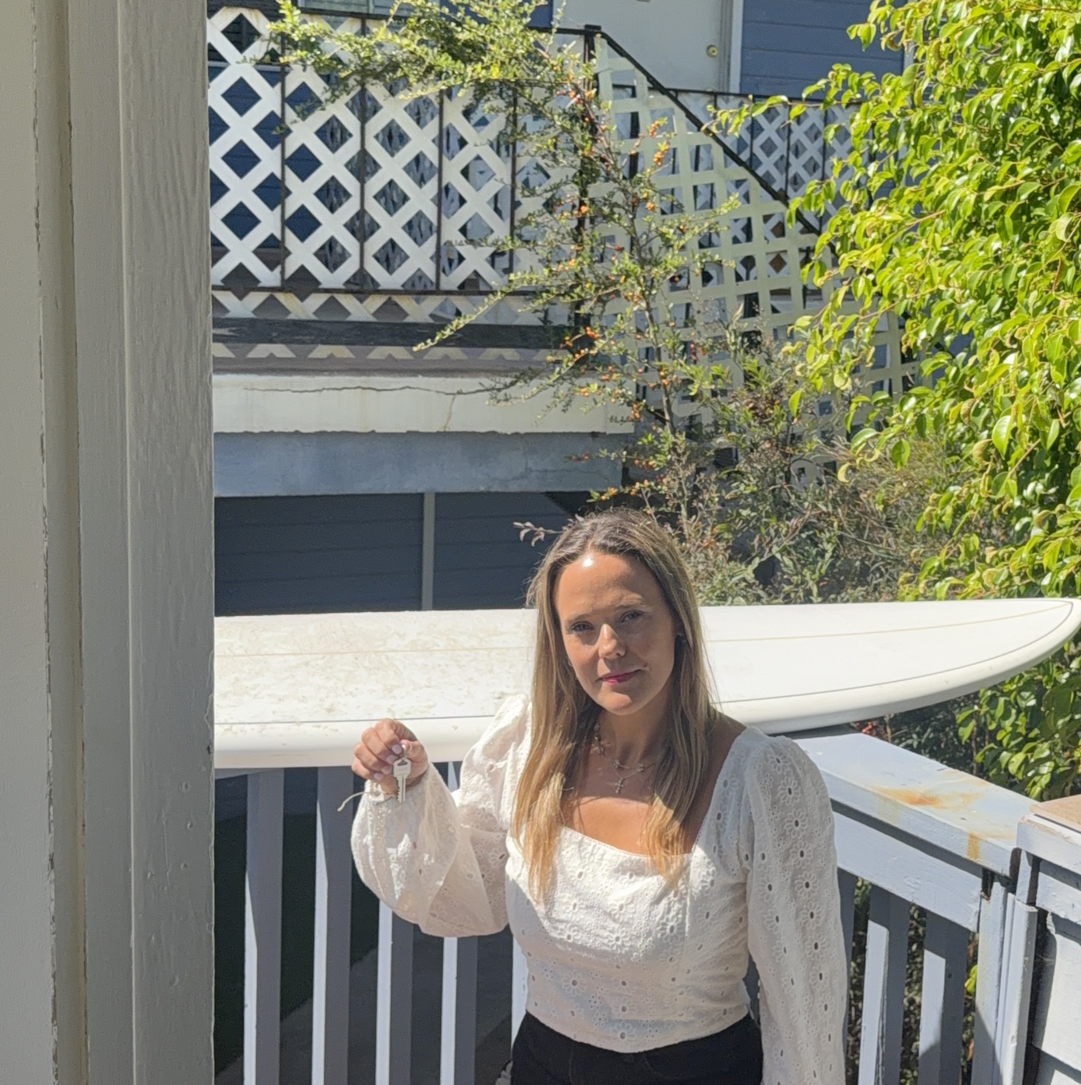 A woman with long blonde hair in a white eyelet blouse holding up keys on a porch deck, with a white surfboard resting behind her on a sunny day in Orange County, California.